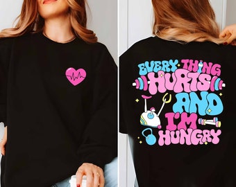 Pode incluir: Camisola de moletom preta com um coração rosa e um gráfico de batimento cardíaco na frente e o texto "EVERYTHING HURTS AND I'M HUNGRY" com um haltere rosa, um kettlebell azul e uma bicicleta de exercício rosa nas costas.