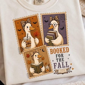 May include: A cream-colored t-shirt featuring a graphic of three postage stamp-style illustrations of ducks wearing bows. The ducks are depicted with autumn leaves, books, and coffee. The text reads BOOKED FOR THE FALL with small stars and hearts.