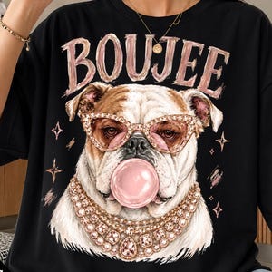 May include: A black t-shirt featuring a graphic of a white bulldog wearing pink cat-eye glasses and a layered gold-toned rhinestone necklace. The dog is blowing a pink bubble gum bubble. The word BOUJEE is printed in a distressed pink font above.