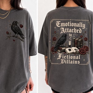 Puede incluir: Una camiseta gris carbón de manga corta con un pequeño gráfico de un cuervo negro sobre una rama de rosa en la parte delantera. La parte trasera muestra un gran gráfico rectangular con un cuervo, libros, rosas y el texto Emotionally Attached to Fictional Villains.