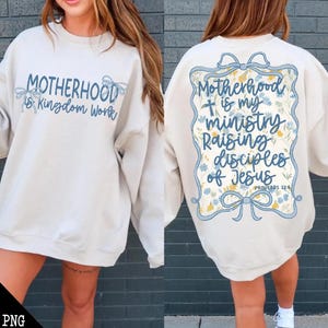 以下が含まれることがあります： 白いスウェットシャツに、青い花の模様と「Motherhood is my ministry Raising disciples of Jesus Proverbs 22:6」という文字が背中に書かれています。スウェットシャツの前には、「Motherhood is Kingdom Work」という文字が青色で書かれています。