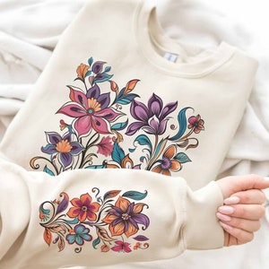 Puede incluir: Una sudadera de color crema con un dise&ntilde;o floral. El dise&ntilde;o presenta una variedad de flores en tonos de rosa, morado, naranja y azul. Las flores est&aacute;n dispuestas en un racimo en la parte delantera de la sudadera y en la manga.