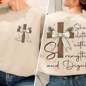 May include: A beige sweatshirt featuring a brown wooden cross adorned with a white bow and green leaves. The text "Proverbs 31:25" is printed beneath the cross. The reverse of the sweatshirt displays the same cross design with the inscription "She is clothed with strength and dignity".