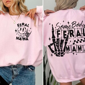 Pu&ograve; includere: Felpa rosa con un design grafico nero che dice "Feral Mama" sul davanti e "Some Body's Feral Mama" sul retro.