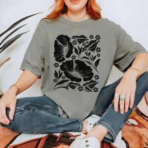 Puede incluir: Una mujer lleva una camiseta verde salvia con un estampado floral negro. La camiseta tiene un corte holgado y mangas cortas. La mujer est&aacute; sentada en una alfombra estampada.