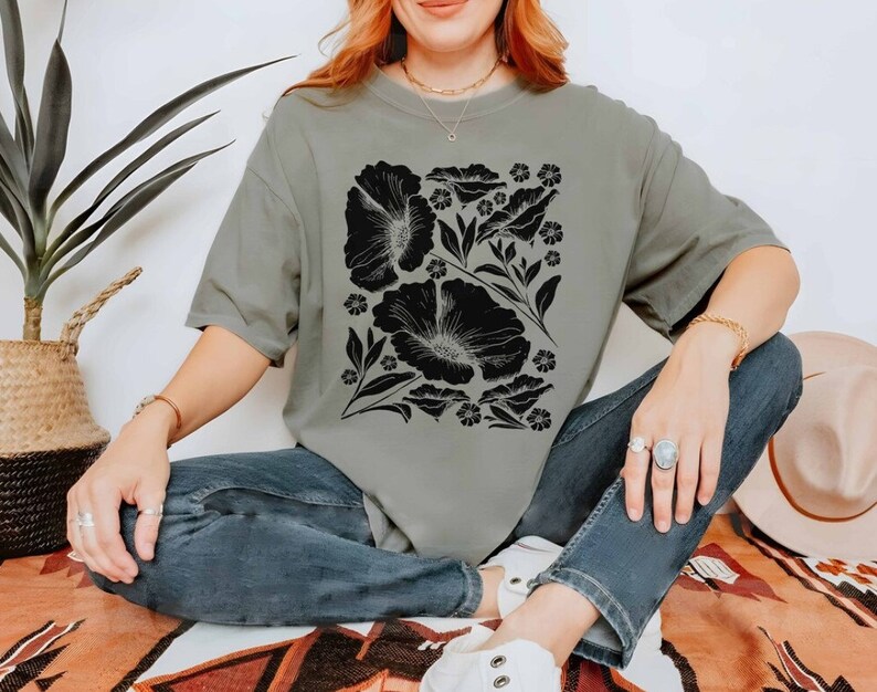 Puede incluir: Una mujer lleva una camiseta verde salvia con un estampado floral negro. La camiseta tiene un corte holgado y mangas cortas. La mujer est&aacute; sentada en una alfombra estampada.