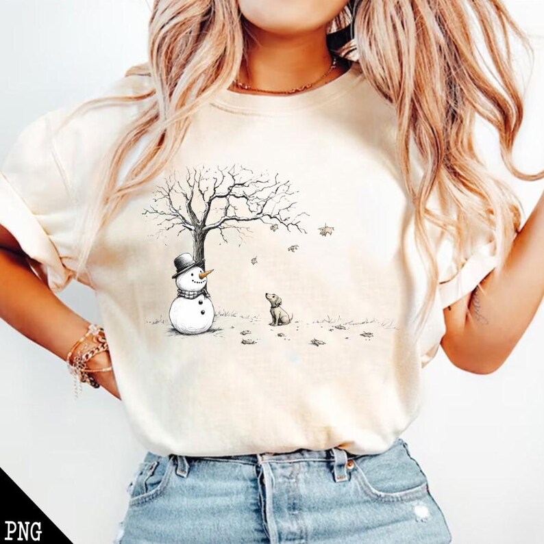 May include: A white t-shirt with a black and white illustration of a snowman and a dog standing under a bare tree with falling leaves.