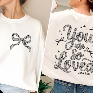 May include: White sweatshirt with a black lace bow on the front and the text "You are so loved" with a black lace design and the text "John 3:16" on the back.