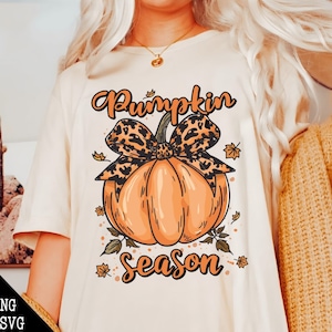 May include: Cream-colored t-shirt featuring a large orange pumpkin with a leopard print bow. The words "Pumpkin Season" are written in orange script. Autumn-themed apparel.