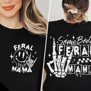 Pu&ograve; includere: Due magliette nere con testo e grafica bianchi. La prima maglietta dice "Feral Mama" con un viso sorridente e una mano scheletrica. La seconda maglietta dice "Some Body's Feral Mama" con una mano scheletrica che fa il segno "Ti amo".