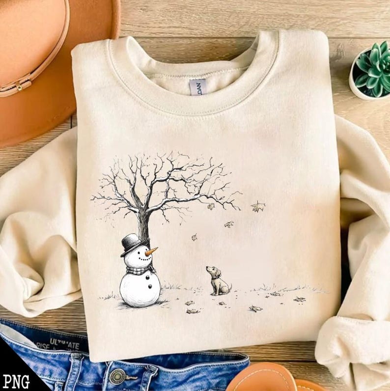 May include: A beige sweatshirt with a black and white illustration of a snowman, a dog, and a bare tree with falling leaves.