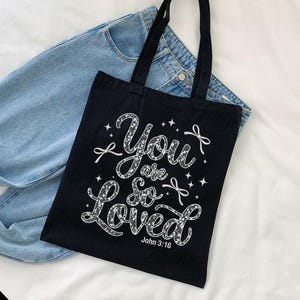 May include: Black tote bag with a white design that says "You are so Loved" with a small bow and stars. The text "John 3:16" is at the bottom of the design.