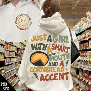 Könnte beinhalten: Weißer Hoodie mit einem Grafikdruck, der "Just a girl with a smart mouth and a cornbread accent" sagt. Die Grafik enthält ein lächelndes Gesicht mit einem Blitz, eine Bratpfanne mit einem Stück Maisbrot und Sterne.