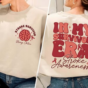 May include: A beige sweatshirt with a pink brain graphic and the text "Stroke Survivor Strong Chloe" on the front. The back of the sweatshirt has the text "In My Survivor Era Stroke Awareness" in pink and red.