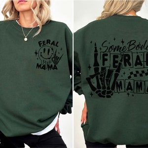 Pu&ograve; includere: Una felpa verde scuro con una stampa grafica nera sul davanti che dice "Feral Mama" con un viso sorridente e una mano scheletrica. La parte posteriore della felpa ha una stampa grafica nera che dice "Some Body's Feral Mama" con una mano scheletrica che fa il segno della pace.