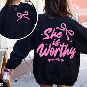 May include: Black sweatshirt with pink lettering that says "She is worthy" with a pink bow above the text and "Proverbs 31" below the text.