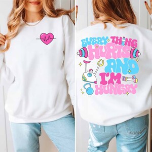 Pu&ograve; includere: Felpa bianca con un cuore rosa e una grafica di battito cardiaco sul davanti e il testo "EVERYTHING HURTS AND I'M HUNGRY" con una cyclette rosa e blu e manubri sul retro.