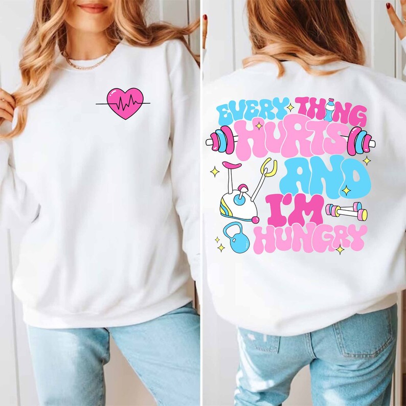 Pu&ograve; includere: Felpa bianca con un cuore rosa e una grafica di battito cardiaco sul davanti e il testo "EVERYTHING HURTS AND I'M HUNGRY" con una cyclette rosa e blu e manubri sul retro.