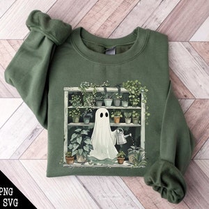 May include: A green sweatshirt with a white ghost watering plants on a shelf. The ghost is holding a watering can and there are many different types of plants on the shelf.