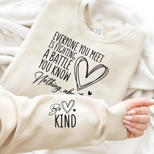 以下が含まれることがあります： 「Everyone you meet is fighting a battle you know. Nothing about... Be Kind」というテキストとハートのグラフィックがプリントされたベージュのトレーナーです。