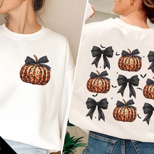 May include: White sweatshirt featuring a leopard print pumpkin with a black bow. The back of the sweatshirt displays a pattern of pumpkins, black bows, and bats. The design is perfect for Halloween or fall.