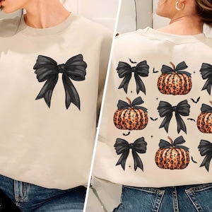 May include: Cream-colored sweatshirt featuring a large black bow graphic on the front. The back of the sweatshirt displays a pattern of leopard print pumpkins and black bows, with bats and stars scattered around.