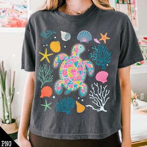 May include: A dark gray cropped t-shirt with a colorful sea turtle design. The turtle is surrounded by coral, seashells, and starfish.