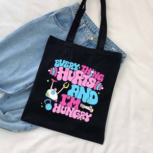 Pu&ograve; includere: Borsa tote nera con una grafica rosa e blu che dice "Tutto fa male e ho fame". La grafica include un manubrio, un kettlebell e un bilanciere.