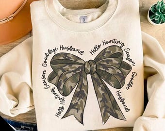 Op de afbeelding: Een beige sweatshirt met een camouflage strik en de tekst "Goodbye Husband Hello Hunting Season" en "Hello Hunting Season Goodbye Husband" rond de strik.