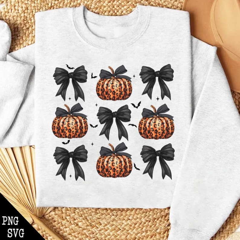 May include: A light gray sweatshirt featuring a repeating pattern of black bows and pumpkins with orange leopard print. Small black bats are scattered around the design, creating a festive Halloween theme.
