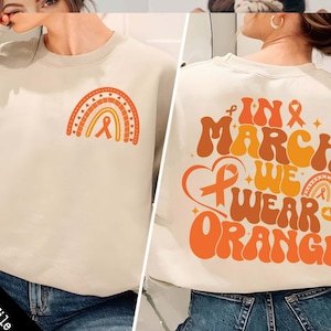 May include: A beige sweatshirt with a rainbow and orange ribbon design on the front and the text "In March We Wear Orange" with an orange ribbon on the back.