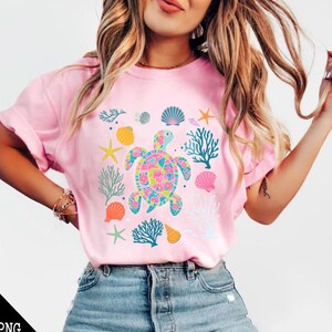 May include: A pink t-shirt with a colorful sea turtle design. The turtle is surrounded by coral, seashells, and starfish.