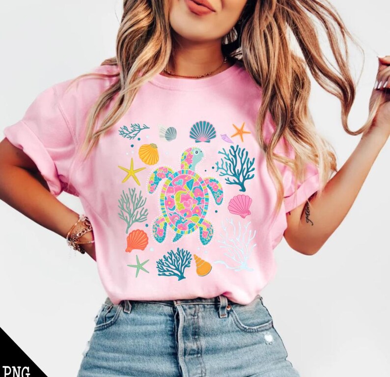 May include: A pink t-shirt with a colorful sea turtle design. The turtle is surrounded by coral, seashells, and starfish.