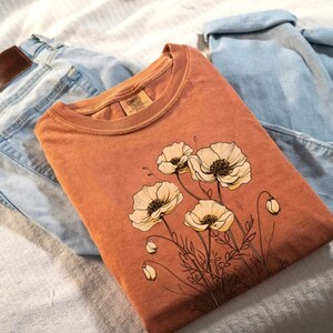 Puede incluir: Una camiseta de color &oacute;xido con un dise&ntilde;o floral con flores de amapola blancas. La camiseta est&aacute; colocada sobre una superficie marr&oacute;n claro con un par de pantalones vaqueros azules.