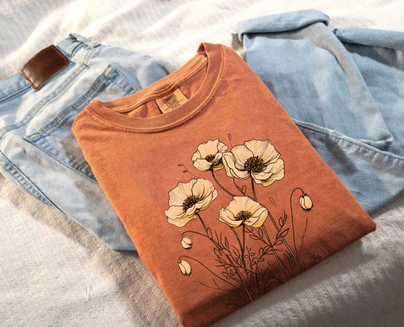 Puede incluir: Una camiseta de color &oacute;xido con un dise&ntilde;o floral con flores de amapola blancas. La camiseta est&aacute; colocada sobre una superficie marr&oacute;n claro con un par de pantalones vaqueros azules.