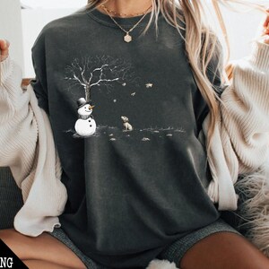 May include: A dark gray sweatshirt with a snowman and a dog graphic. The snowman is wearing a black hat and a scarf. The dog is sitting and looking at the snowman. The graphic is surrounded by falling leaves.