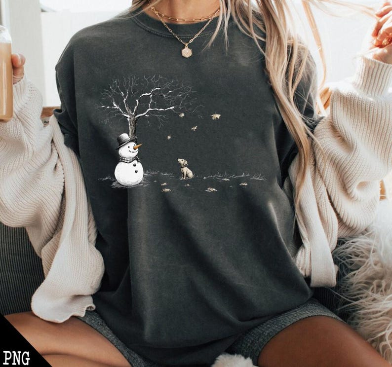 May include: A dark gray sweatshirt with a snowman and a dog graphic. The snowman is wearing a black hat and a scarf. The dog is sitting and looking at the snowman. The graphic is surrounded by falling leaves.