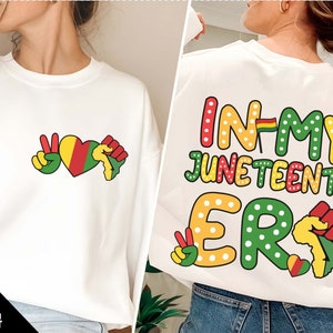 May include: White sweatshirt with a peace sign, heart, and fist graphic on the front and the text "In My Juneteenth Er" on the back. The graphic features red, yellow, and green colors.