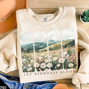 Op de afbeelding: Een beige sweatshirt met een aquarelprint van een bloemenveld en bergen. De tekst "Let Kindness Bloom" is onder de afbeelding gedrukt.