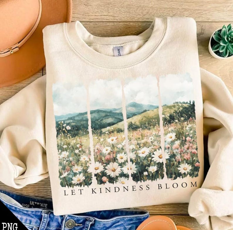 Op de afbeelding: Een beige sweatshirt met een aquarelprint van een bloemenveld en bergen. De tekst "Let Kindness Bloom" is onder de afbeelding gedrukt.