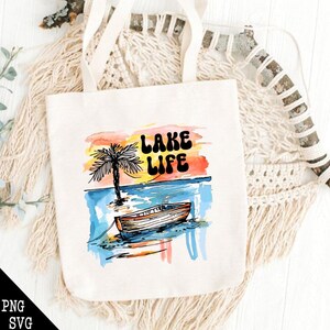 May include: A white tote bag with a watercolor design of a palm tree, a boat, and a sunset. The text "LAKE LIFE" is written in black letters on the bag.