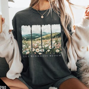 Op de afbeelding: Een donkergrijs T-shirt met een bloemenprint en de tekst "Let Kindness Bloom". De print toont een landschap in aquarelstijl met bergen en bloemen.