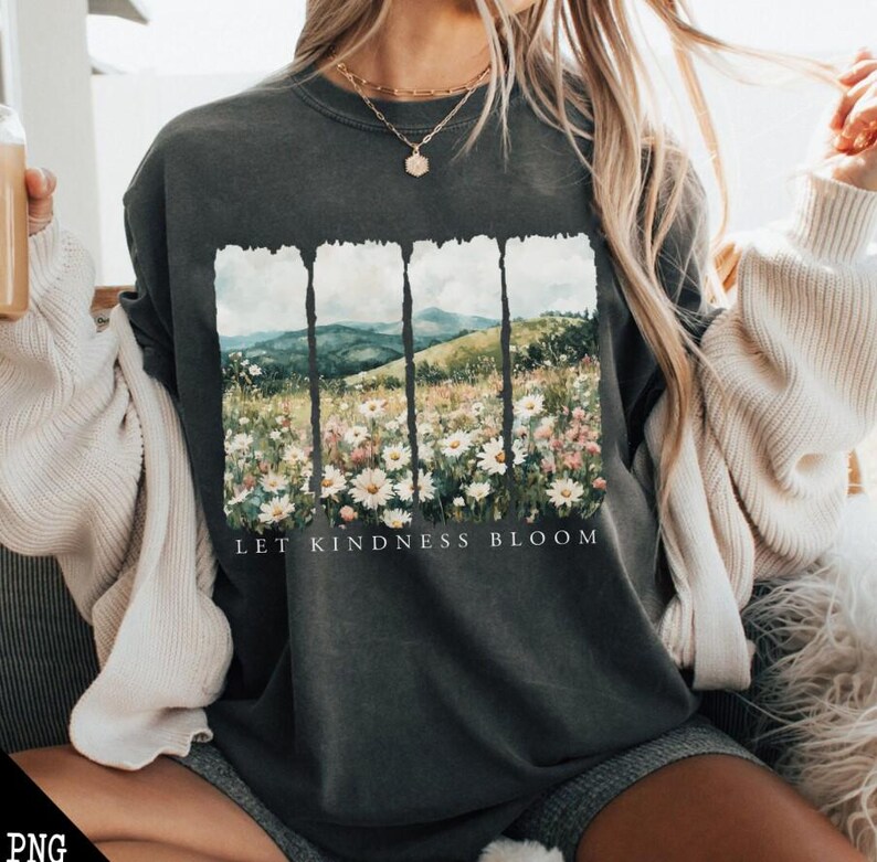 Op de afbeelding: Een donkergrijs T-shirt met een bloemenprint en de tekst "Let Kindness Bloom". De print toont een landschap in aquarelstijl met bergen en bloemen.
