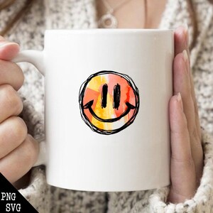 May include: White ceramic mug with a hand-drawn smiley face in orange, yellow, and red colors. The smiley face is outlined in black.