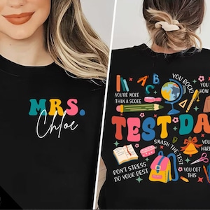 May include: A black t-shirt with a colorful design featuring the text "MRS. Chloe" on the front and "TEST DAY" with colorful school-themed illustrations on the back.