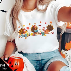 May include: White t-shirt with a graphic of three turkeys reading books. The turkeys are wearing blue shirts and have brown feathers. The background is white with orange and yellow leaves.