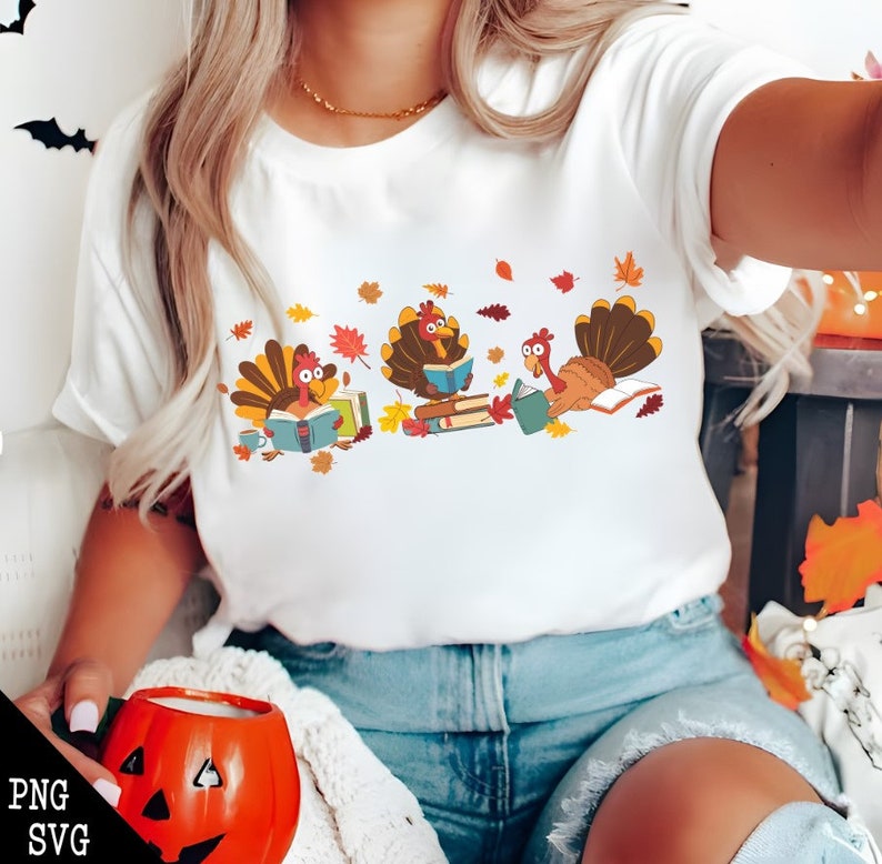 May include: White t-shirt with a graphic of three turkeys reading books. The turkeys are wearing blue shirts and have brown feathers. The background is white with orange and yellow leaves.