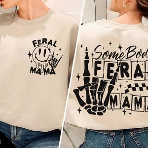 Pu&ograve; includere: Una felpa beige con un design grafico nero sul davanti che dice "Feral Mama" con un viso sorridente e una mano scheletrica. Il retro della felpa ha un design grafico nero che dice "Somebody's Feral Mama" con una mano scheletrica e uno schema a scacchi.