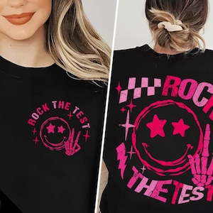 May include: Black t-shirt with a pink graphic of a smiley face with stars for eyes and the text "Rock the Test" in a pink, distressed font.