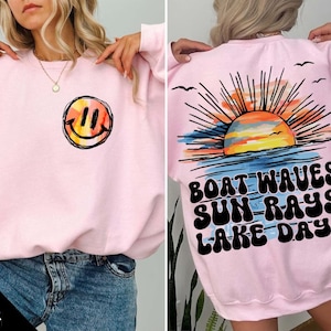May include: Pink sweatshirt with a smiley face on the front and a sunset graphic with the text "Boat Waves Sun Rays Lake Days" on the back.